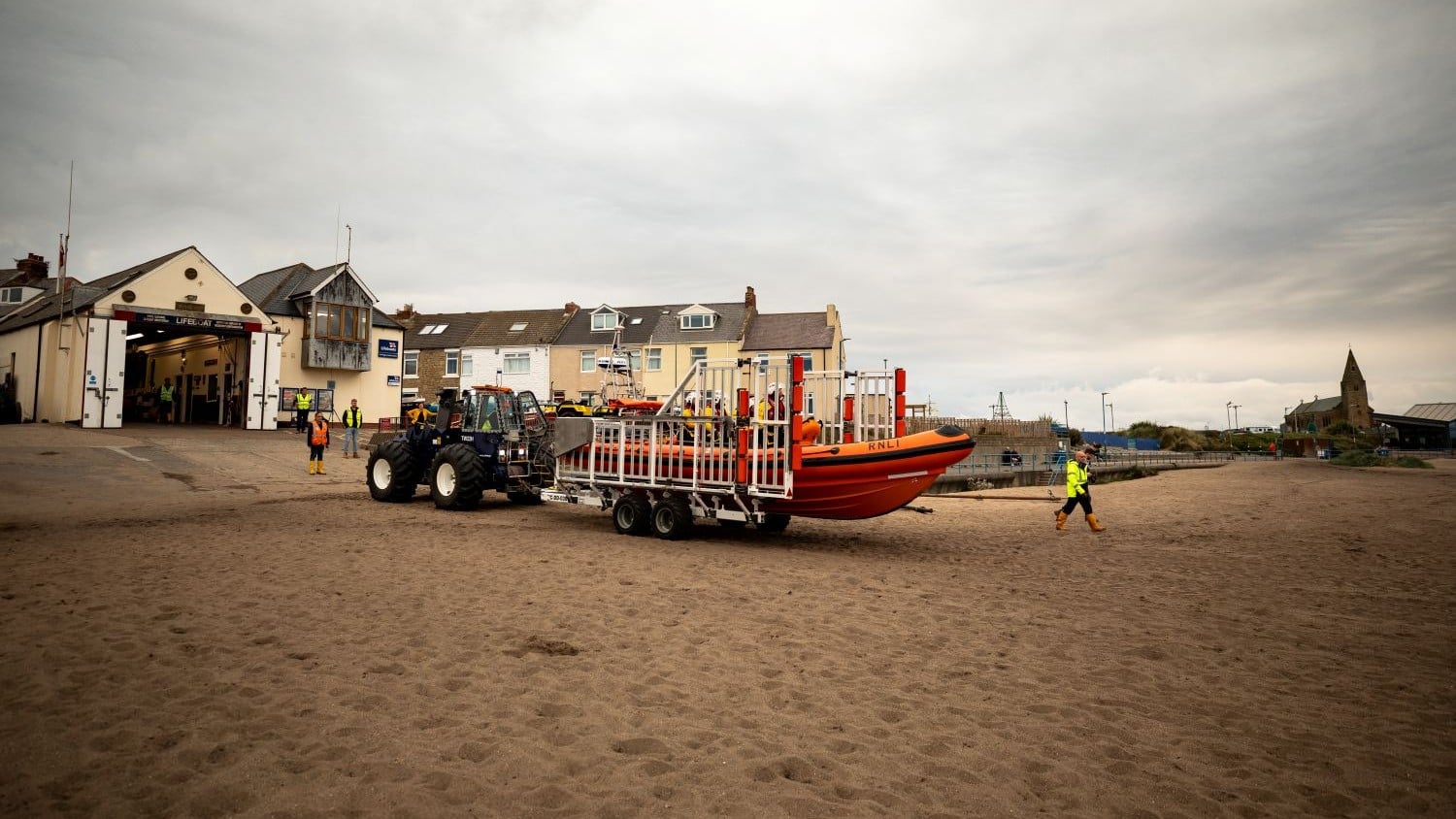 A one storey cream building with pointy roof whose doors open onto a beach. A tractor is pushing a rig with an ILB on it.