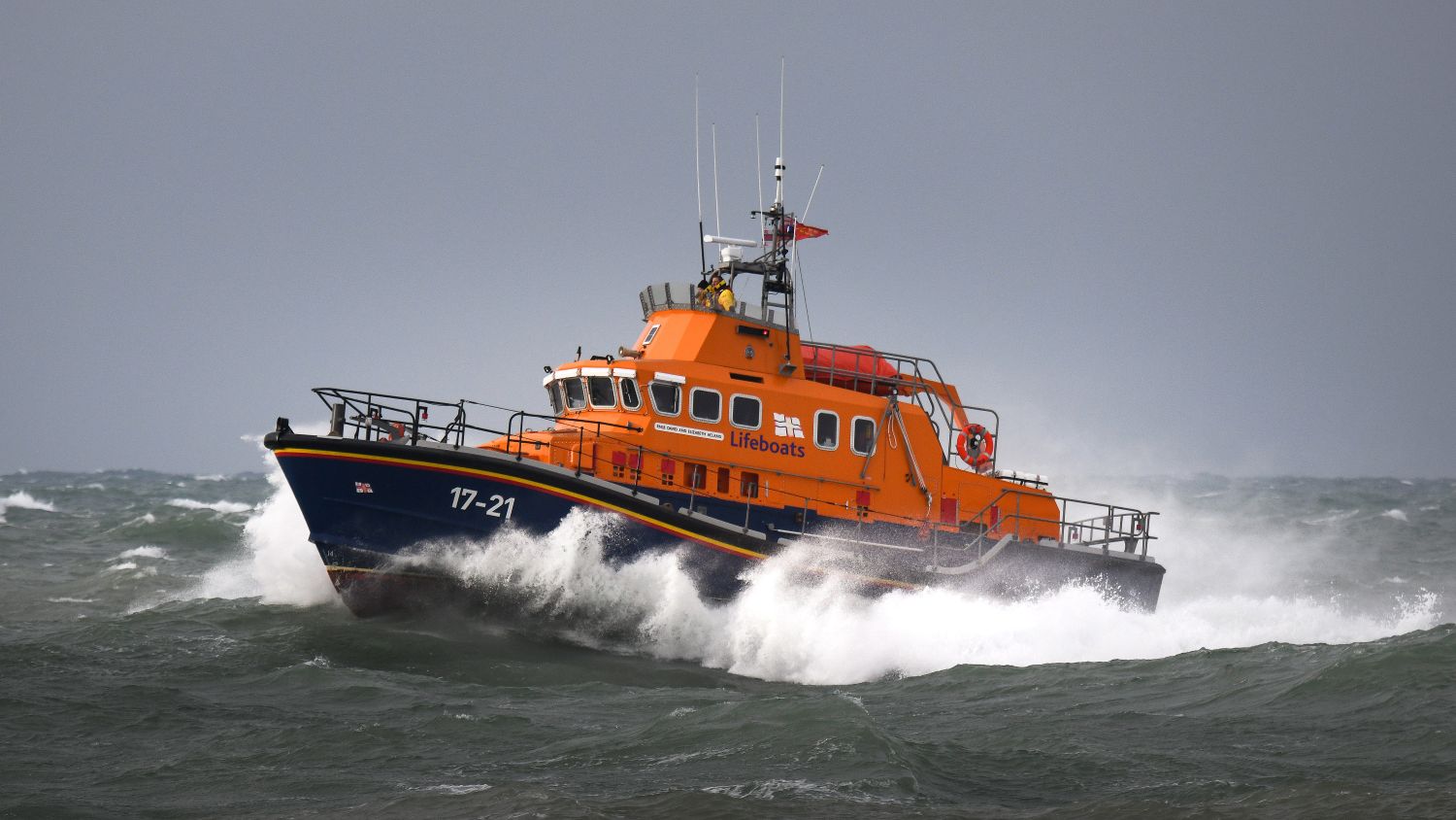 Orange RNLI lifeboat (17‑21) cutting through rough, choppy sea with waves crashing around it under a grey, overcast sky