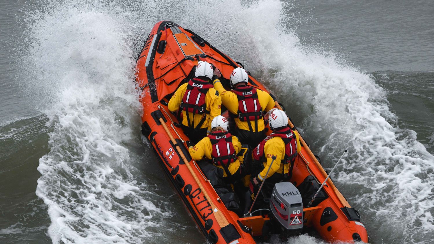 RNLI lifeboat with five crew in helmets powers through rough grey waves, surrounded by heavy spray.