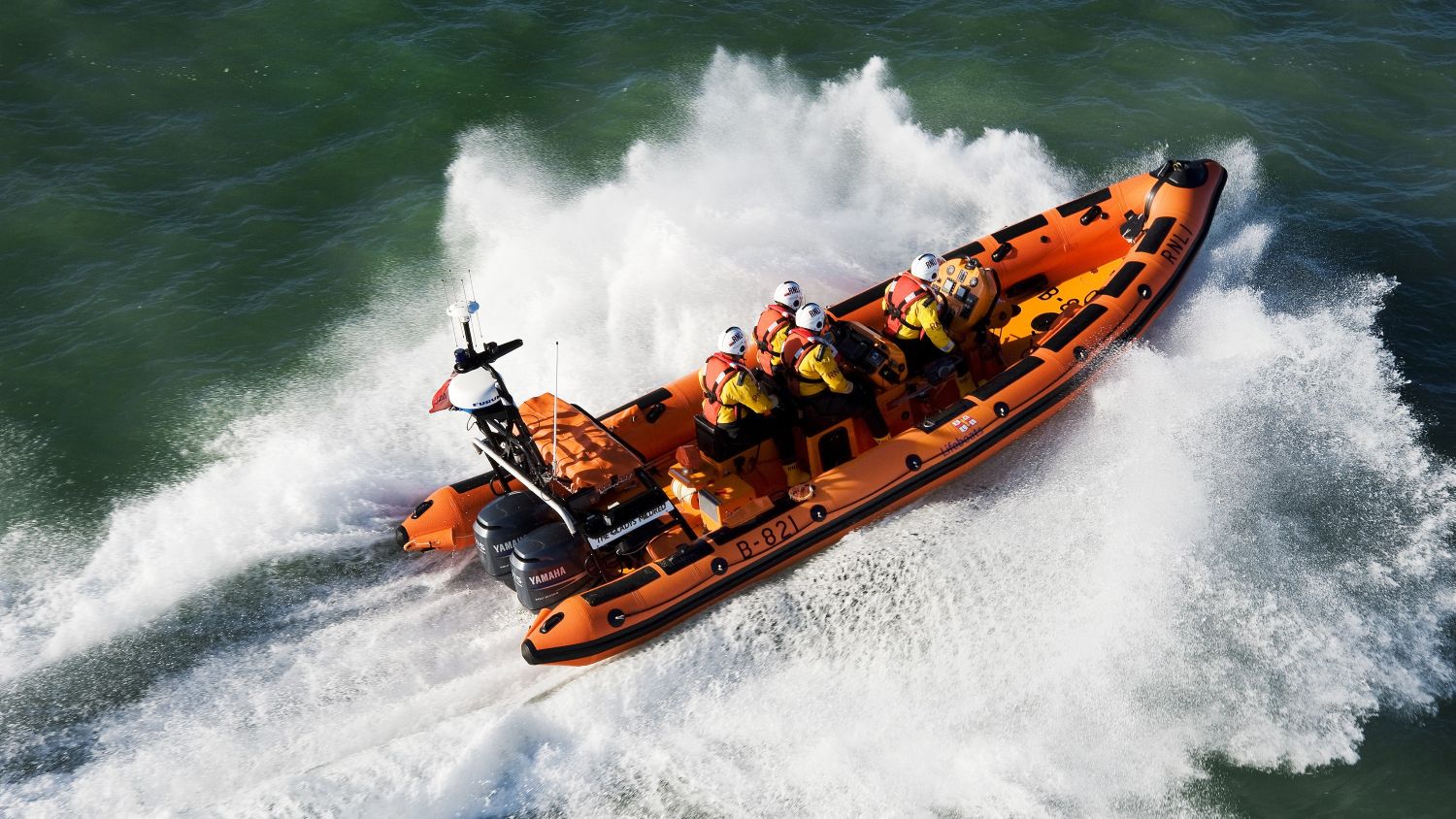 RNLI lifeboat speeds across turquoise water, crew braced as the boat throws up large white spray.