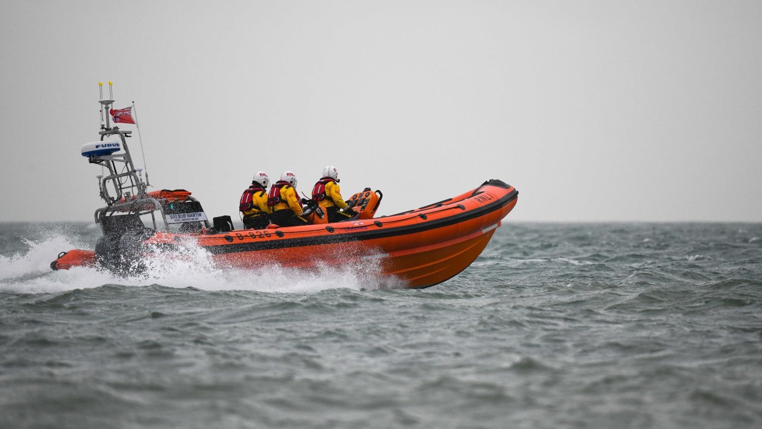 An orange lifeboat speeds across choppy grey water, carrying four crew members in helmets and high-visibility jackets under an overcast sky.