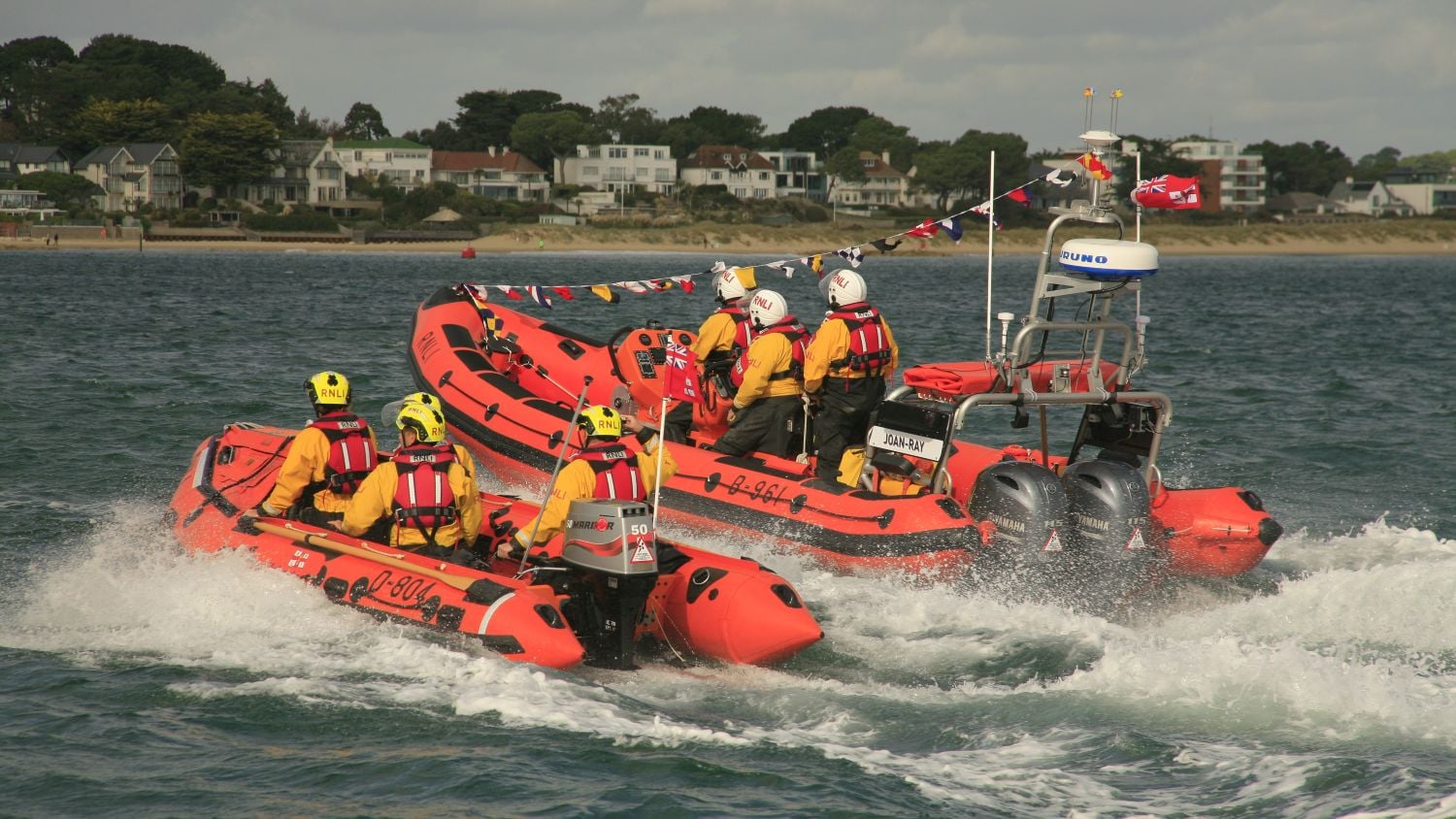 RNLI D class lifeboat Gladys-Maud in the foreground and Atlantic 85 lifeboat Joan-Ray in the background, with the shoreline and houses visible behind. Joan-Ray is decorated with bunting