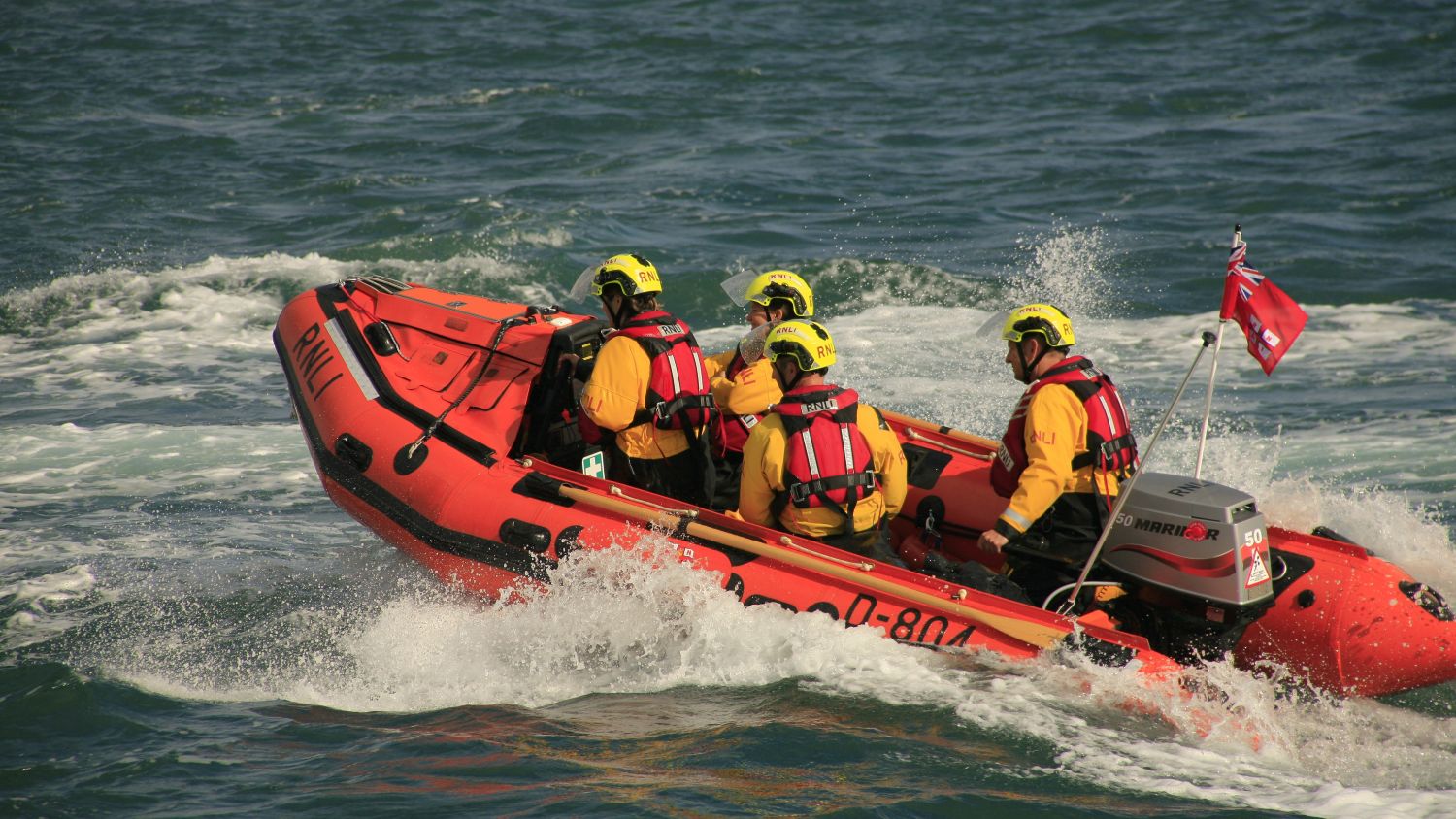 RNLI D class lifeboat Gladys-Maud travelling at speed across the water with crew onboard wearing yellow helmets and lifejackets.