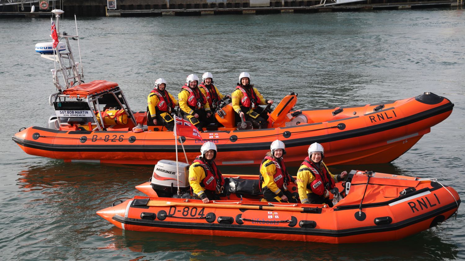 Two orange RNLI lifeboats on the water, each carrying crew members wearing helmets and lifejackets, with harbour buildings in the background.