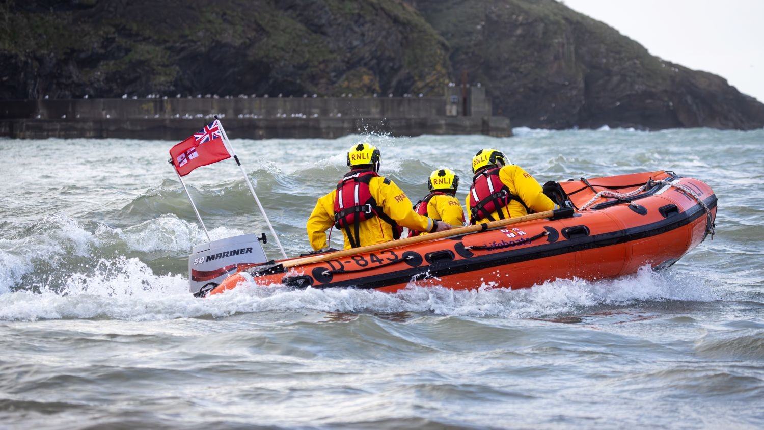 An RNLI RIB powers through choppy coastal waters, with three crew members in yellow helmets and waterproof jackets onboard, approaching a harbour backed by rocky cliffs.
