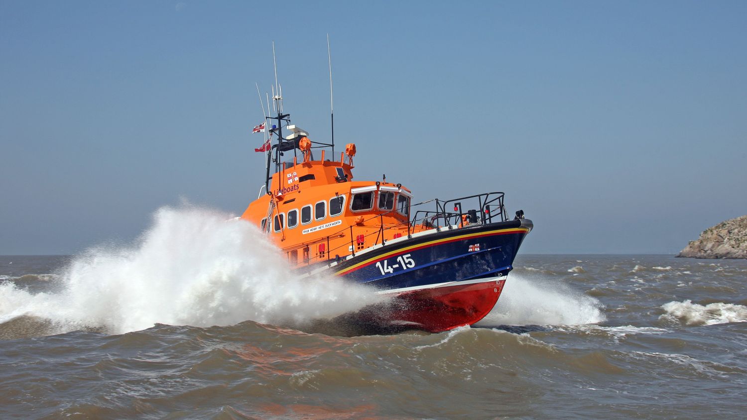 A lifeboat powering through rough, choppy waves, throwing up white spray as it speeds across open water. Its number 14-15 is visible.