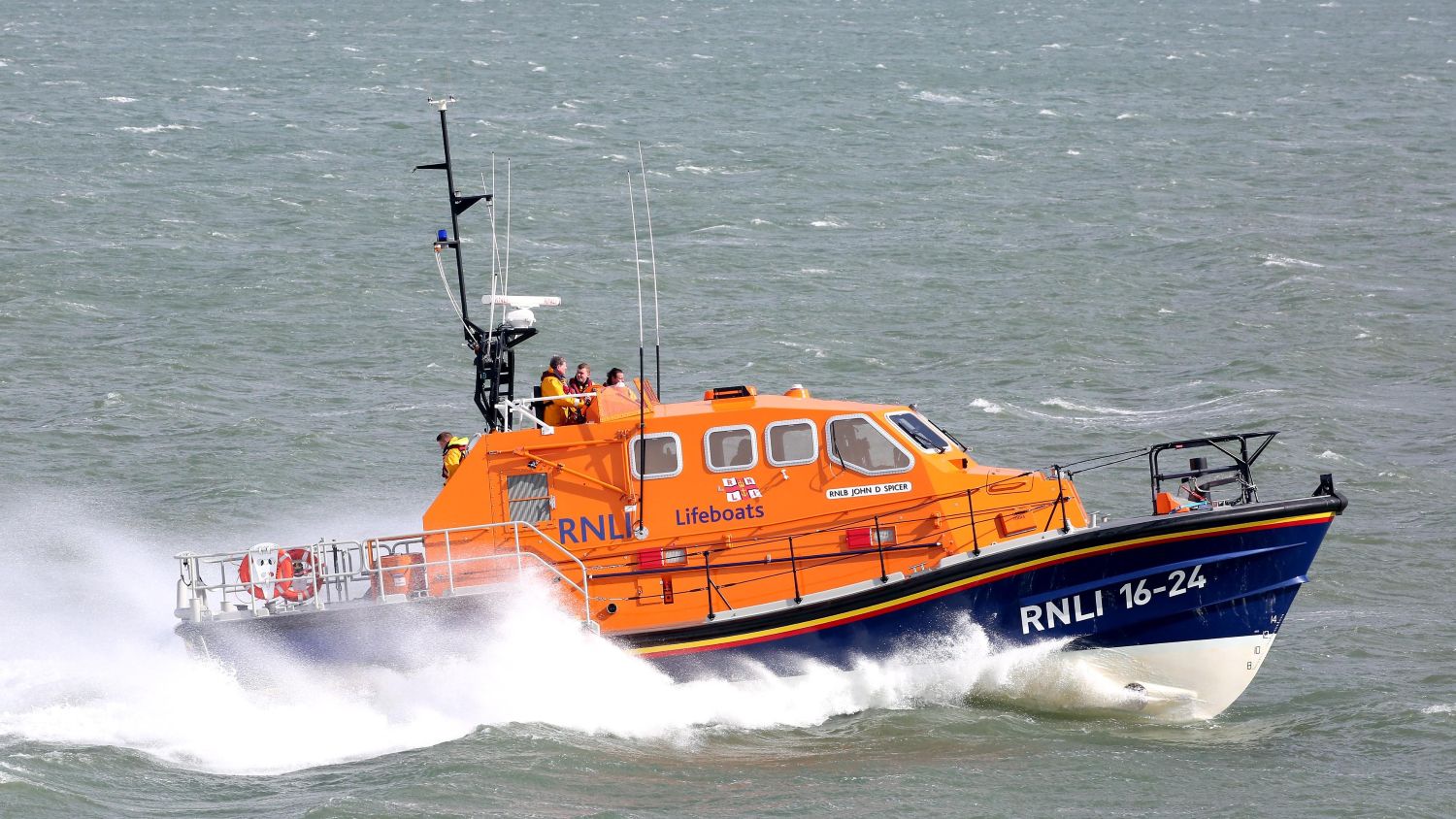 RNLI lifeboat speeding through choppy seas, carrying four crew members in high‑visibility yellow helmets and protective gear as waves splash around the orange boat.