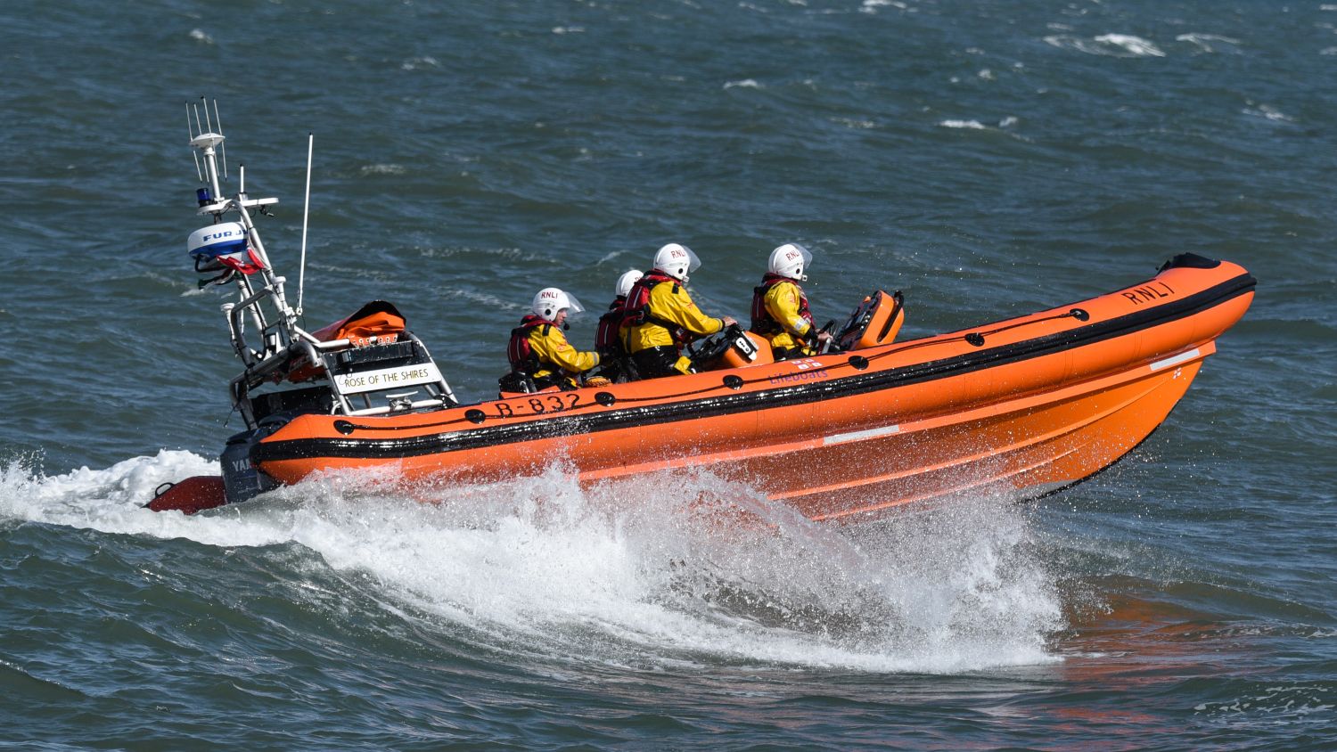 RNLI lifeboat powering through rough seas, with four crew members in high‑visibility yellow helmets and waterproof gear as waves break around the orange boat.
