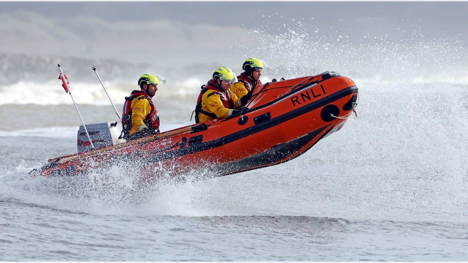 RNLI lifeboat moving through rough seas, with four crew members in yellow helmets and waterproof gear as waves crash around the boat.