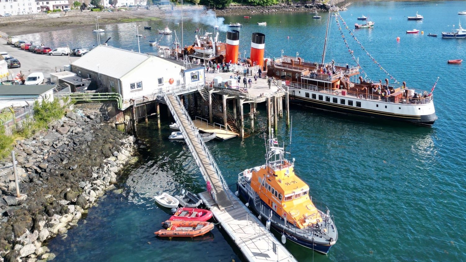 A one storey white building with a steep gangway down to a pontoon where an all weather lifeboat is moored. There is a paddle steamer pulled up on the opposite side of the quay to the station.