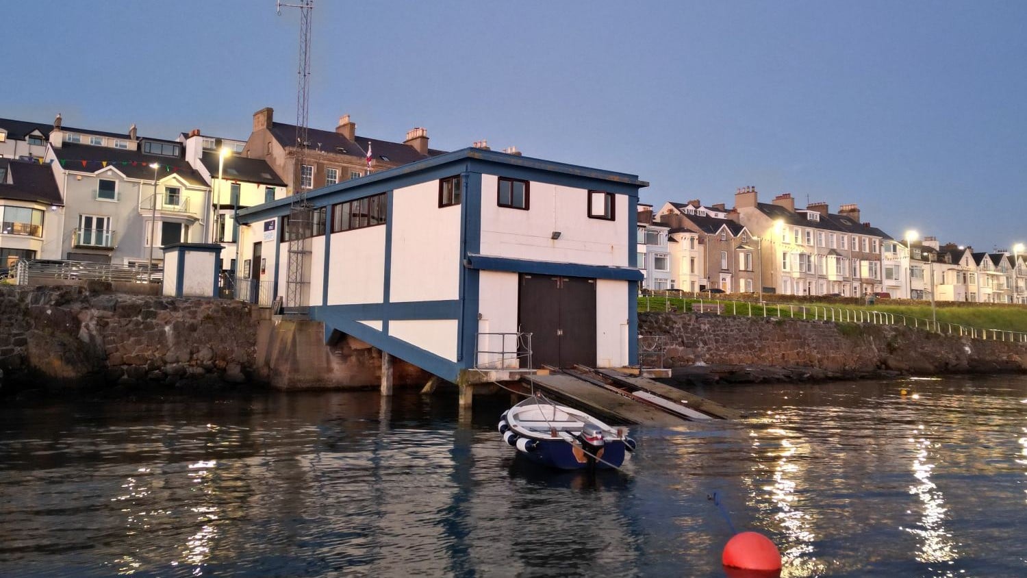 A white and blue building with a flat roof that extends on stilts from the waters edge with a slipway.