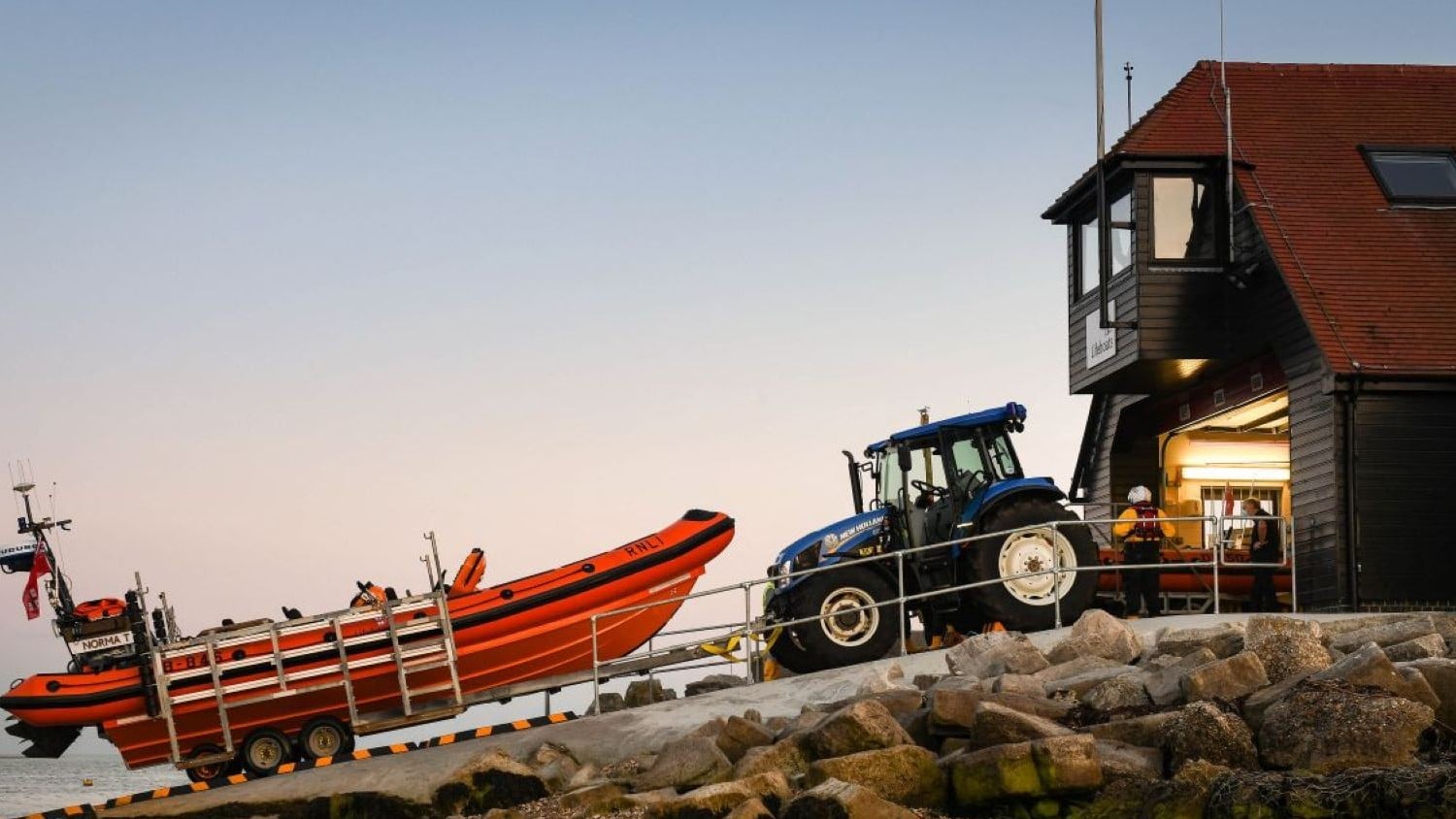 A two storey dark wood building with sloping red tile roof. A tractor and B class lifeboat sit on the slipway.