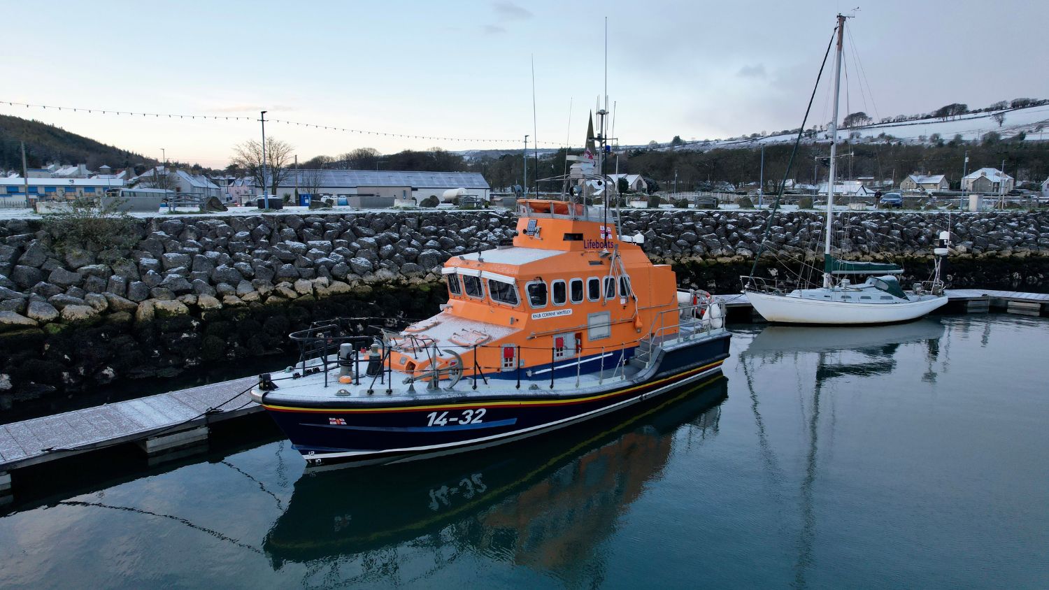 A Red Bay Trent class RNLI lifeboat, numbered 14-32, moored at a calm harbour beside a rocky breakwater. The orange and blue vessel reflects on the still water, with a sailing boat and snow-dusted hills visible in the background.