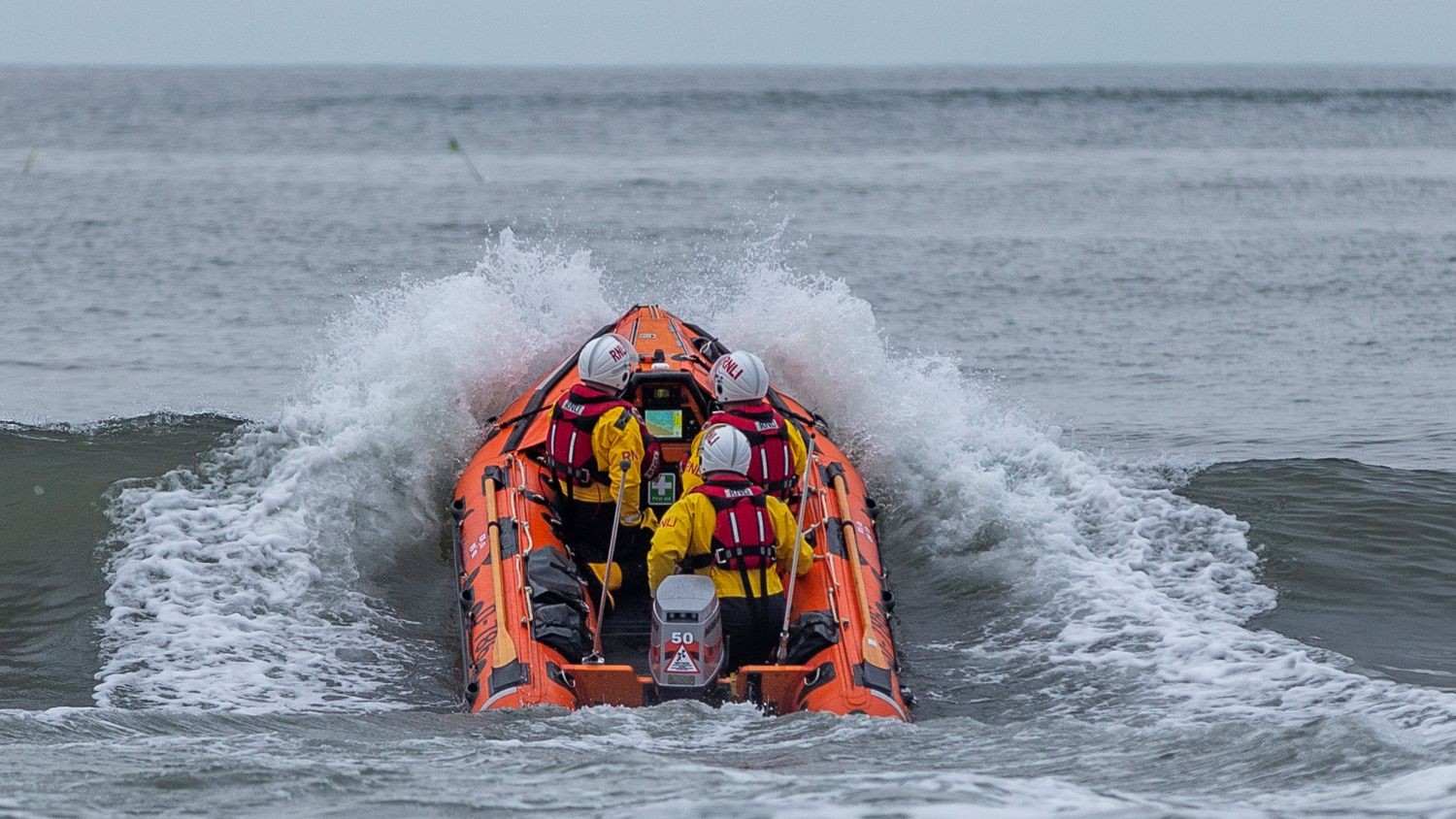 A small rib with three crew crests a grey wave, with lots of white spray.