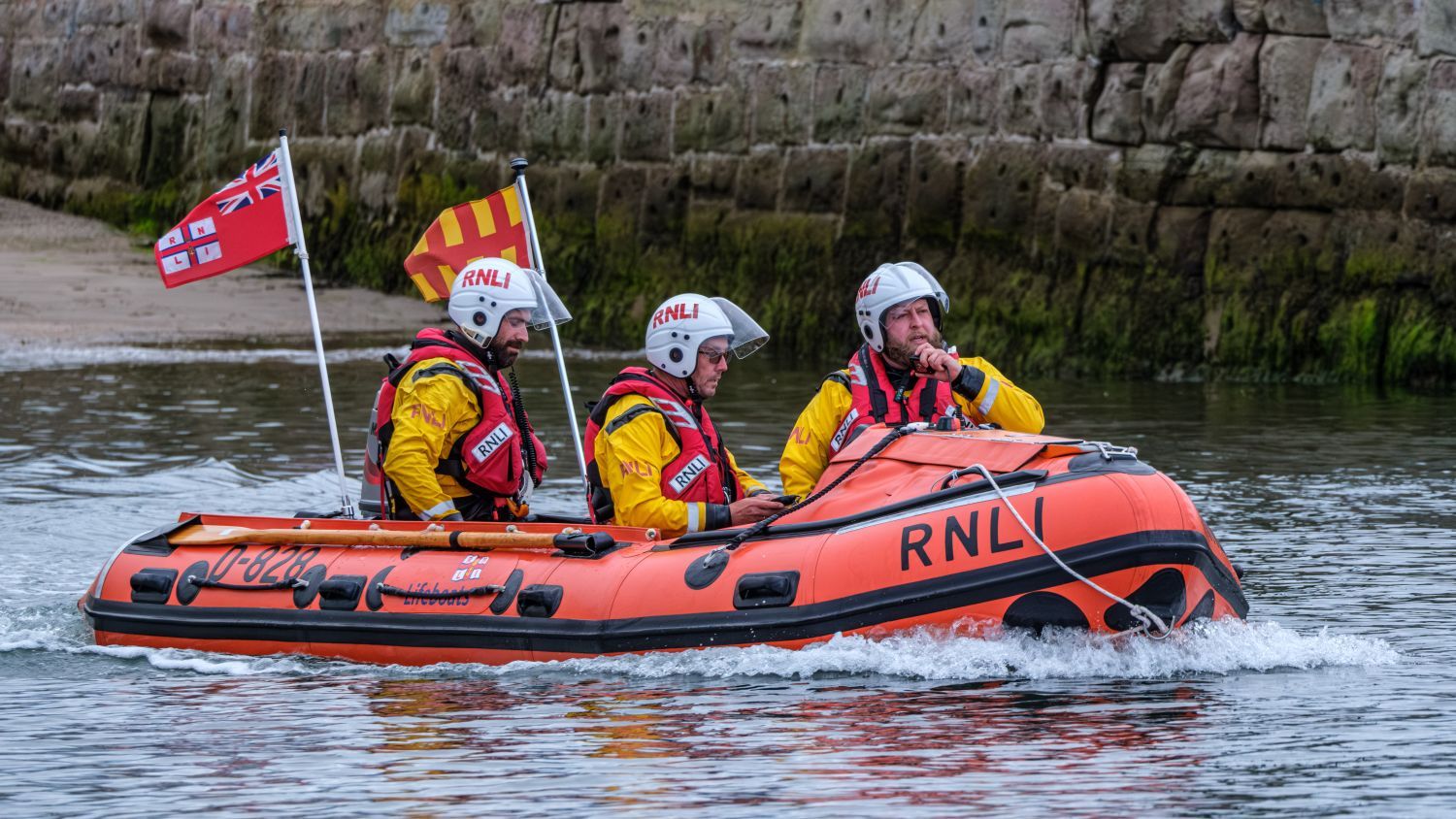Three RNLI volunteers wearing helmets and lifejackets travel in an orange inflatable lifeboat, flying RNLI flags near a stone harbour wall.