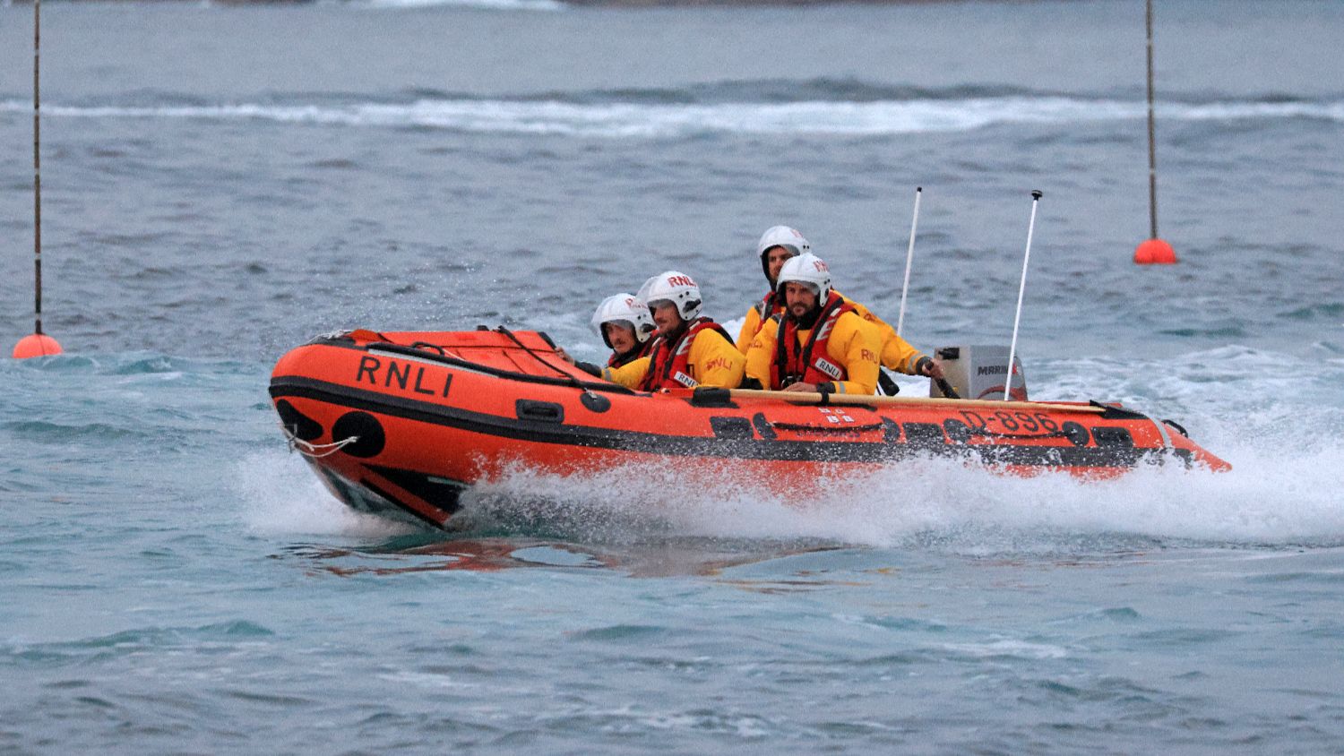 RNLI D‑class inflatable lifeboat travelling at speed across choppy water, carrying four helmeted crew members in yellow protective clothing, with spray breaking along the bow and marker buoys visible in the background.