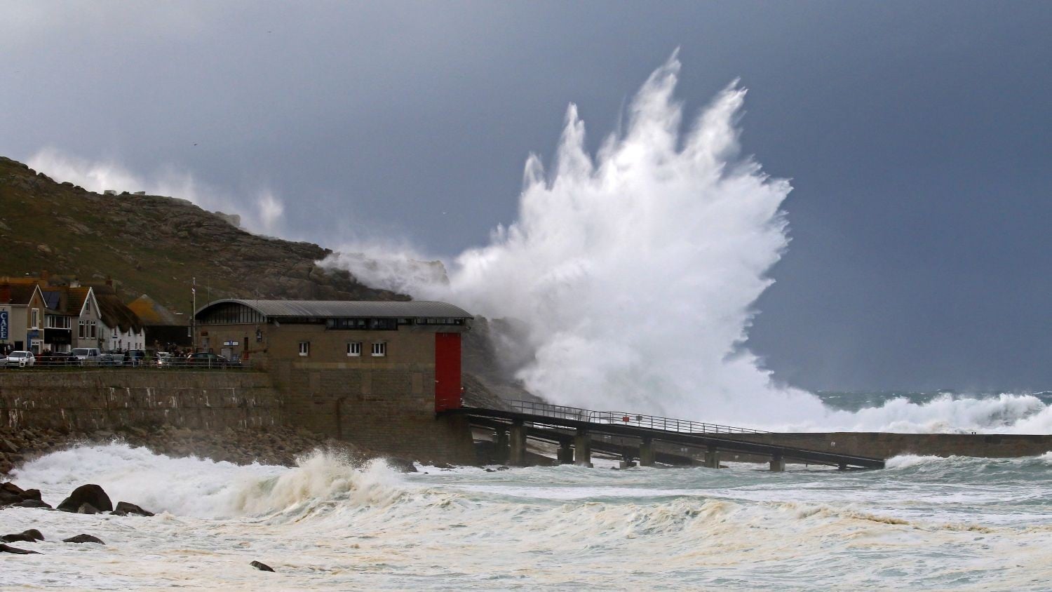 A brown building with curved roof and a long slipway. A huge wave breaks on the headland behind the station.