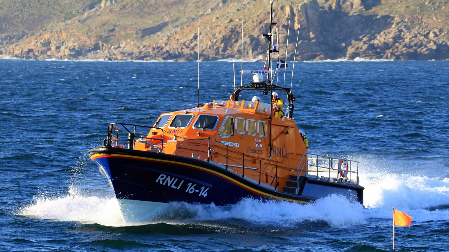 RNLI Tamar‑class lifeboat RNLI 16‑14 moving through rolling waves, with a rocky coastline in the background and water spraying along the hull.