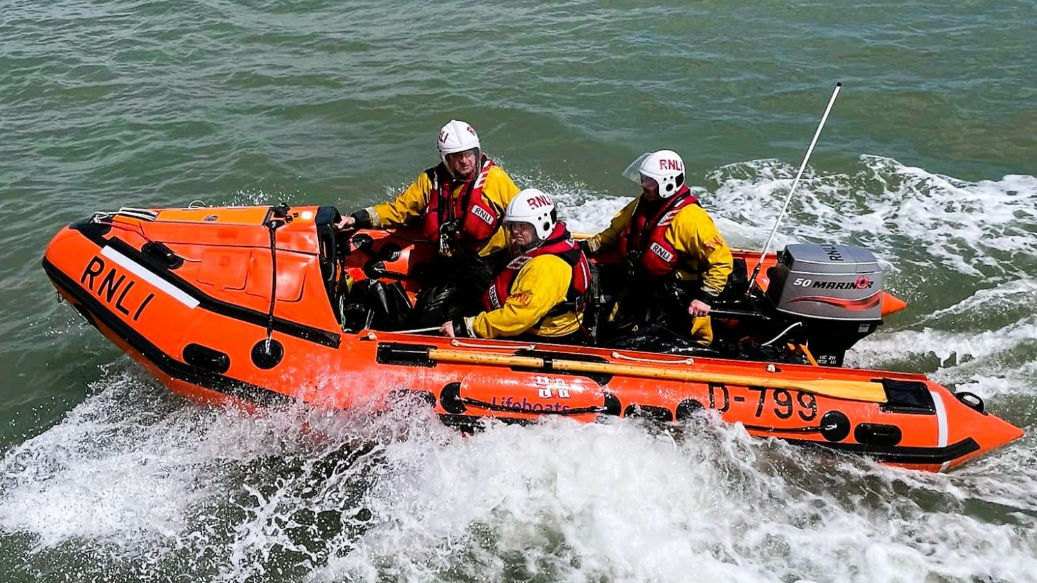 RNLI D‑class inflatable lifeboat travelling at speed through choppy water, with three helmeted crew members in yellow protective clothing as white water breaks along the hull.
