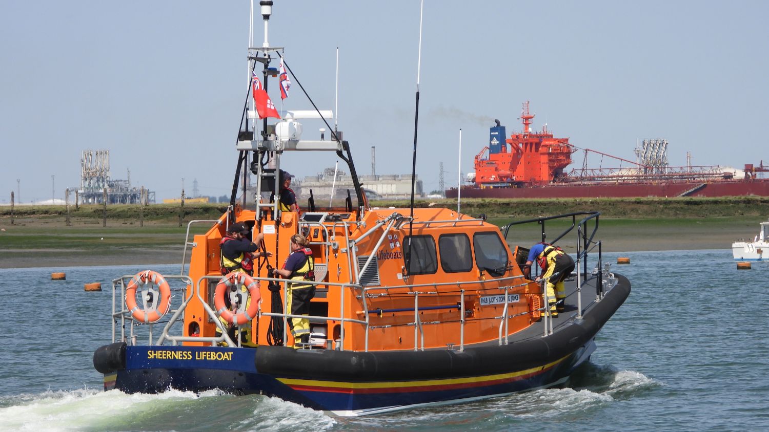 Orange RNLI lifeboat underway on calm water, viewed from behind, with several crew members on deck as it leaves harbour, industrial shoreline and a large red cargo ship visible in the background.