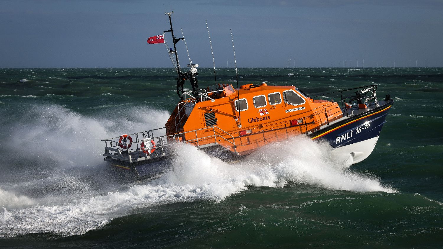 Orange RNLI all‑weather lifeboat RNLI 13‑15 powering through rough, dark green seas, throwing up heavy spray alongside the vessel.