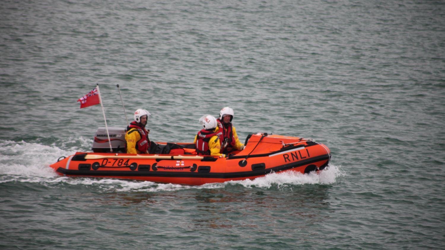 RNLI D‑class inflatable lifeboat carrying three helmeted crew members, travelling steadily across calm water.