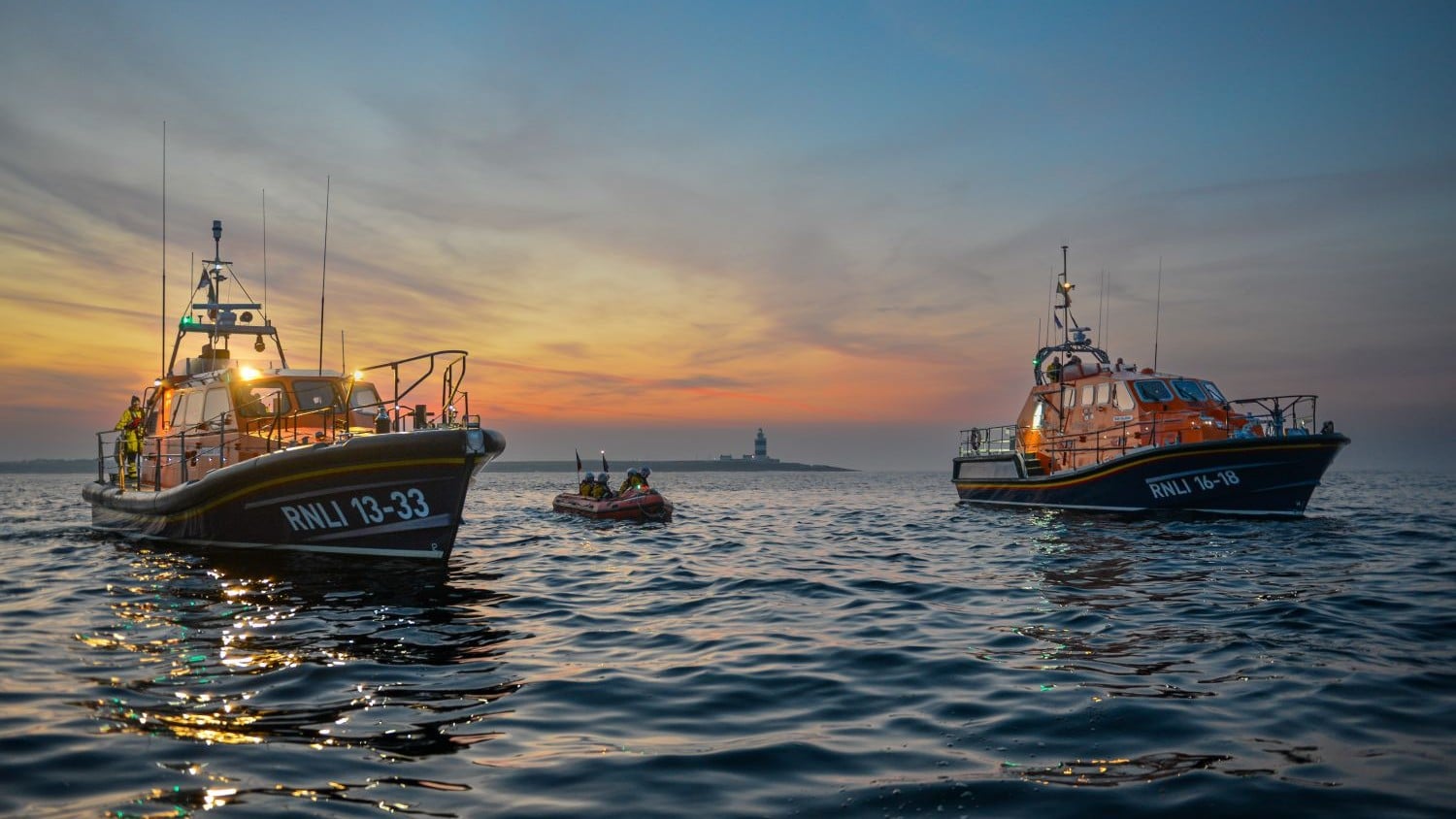 Three different classes of lifeboat gather on a calm sea at sunrise.