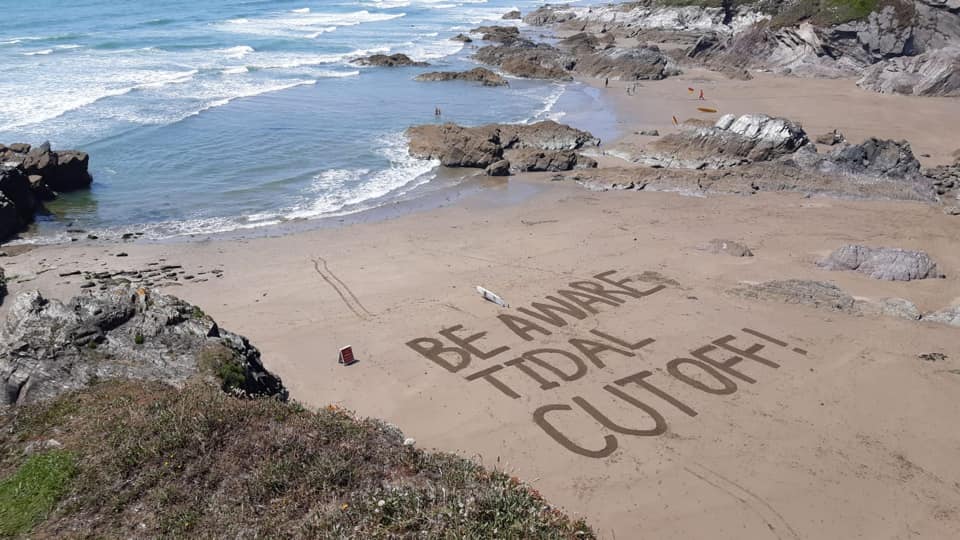 Sand art on a beach which says 'Be aware tidal cut off' in large letters.