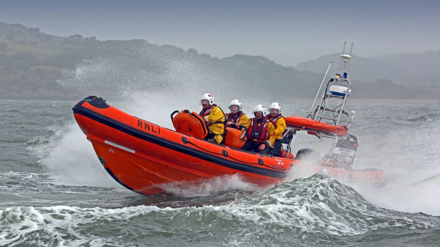 RNLI D‑class inflatable lifeboat launching over rough seas, with four helmeted crew members in yellow protective clothing as the bow lifts over a wave and heavy spray rises around the boat, with a hazy coastline in the background.
