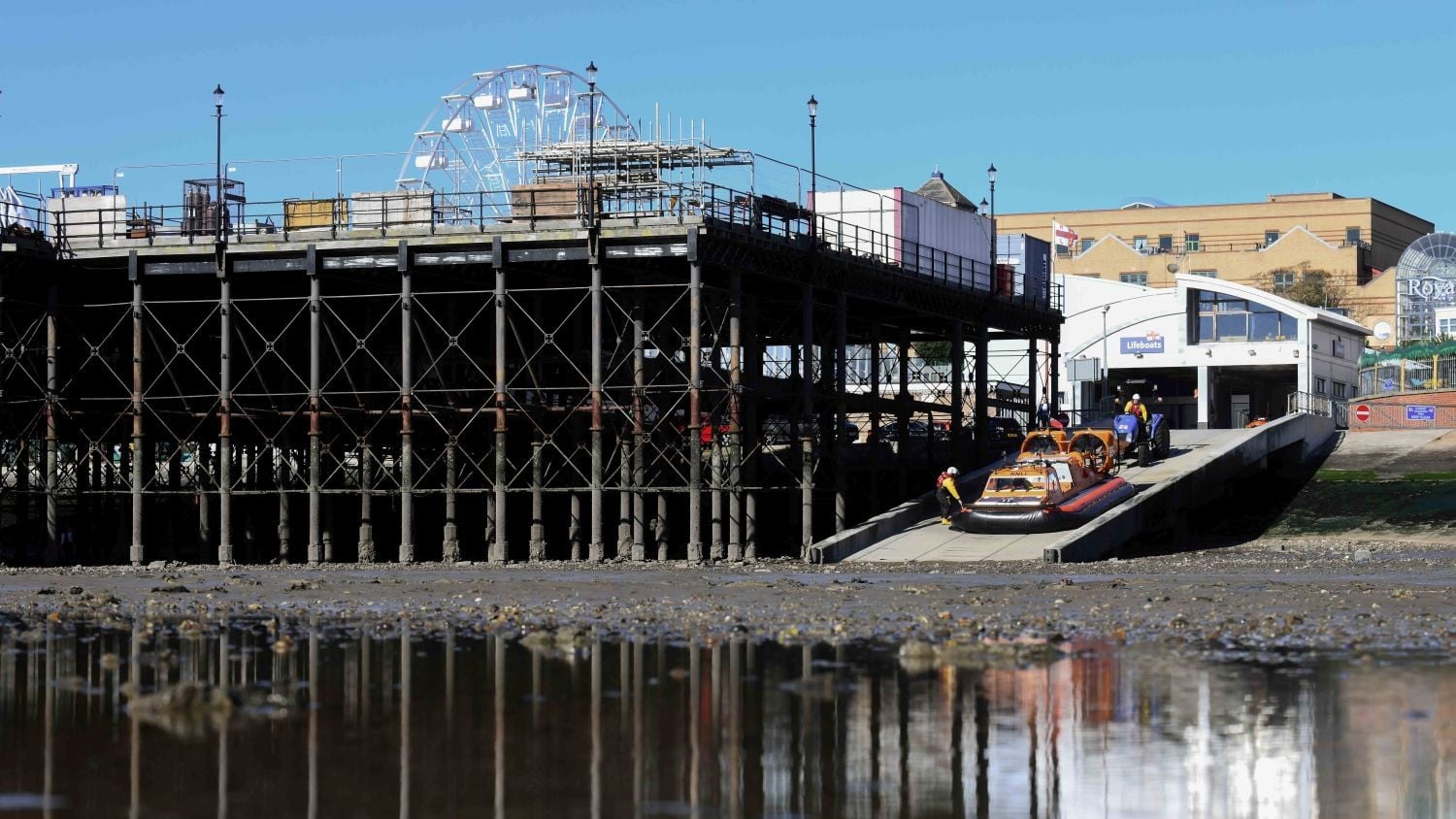 A two-storey white building with curved rooves next to the tall supports for a pier. There is a hovercraft on the slipway.