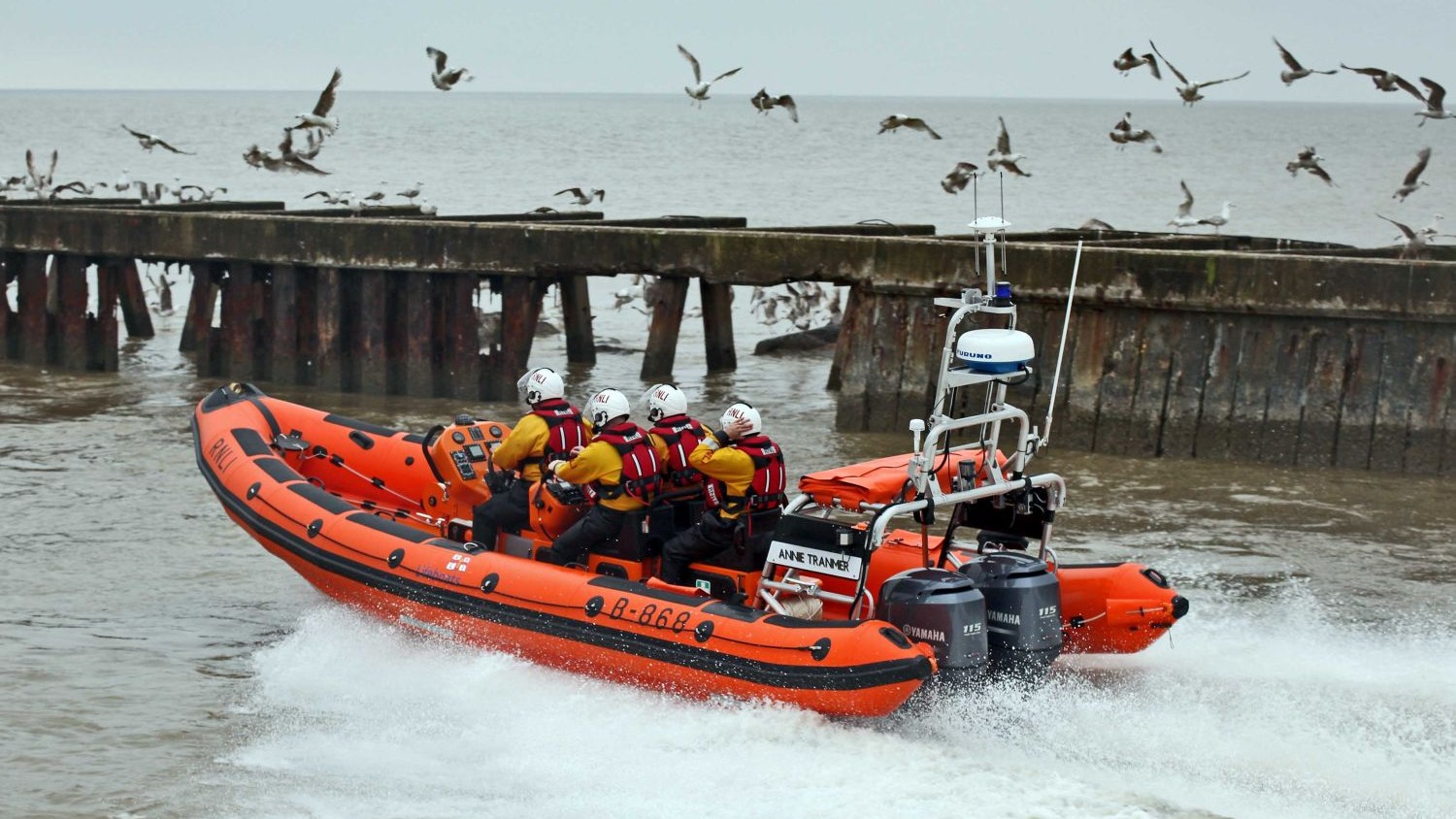 A large rib with four crew passes along a pier with gulls flying around it.