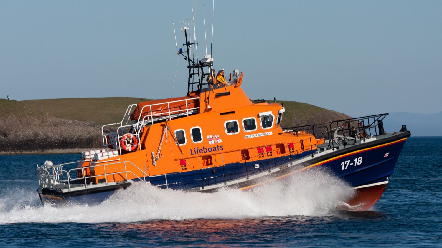 Orange RNLI all‑weather lifeboat RNLI 17‑18 travelling at speed across calm blue water, viewed from the side, with white spray breaking along the bow and low, grassy islands visible in the background.