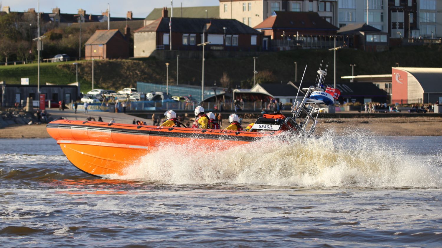 RNLI inshore lifeboat travelling at speed close to shore, with crew members onboard as white spray rises from the bow, beachfront buildings and promenade visible in the background.