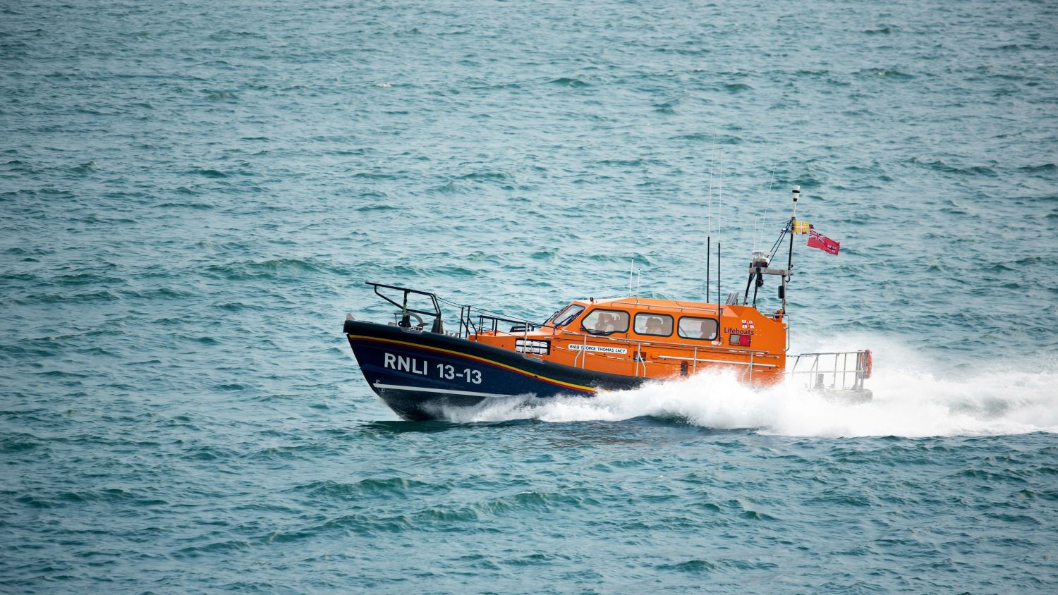 An Shannon class all weather lifeboat powers across blue sea creating white spray in its wake.