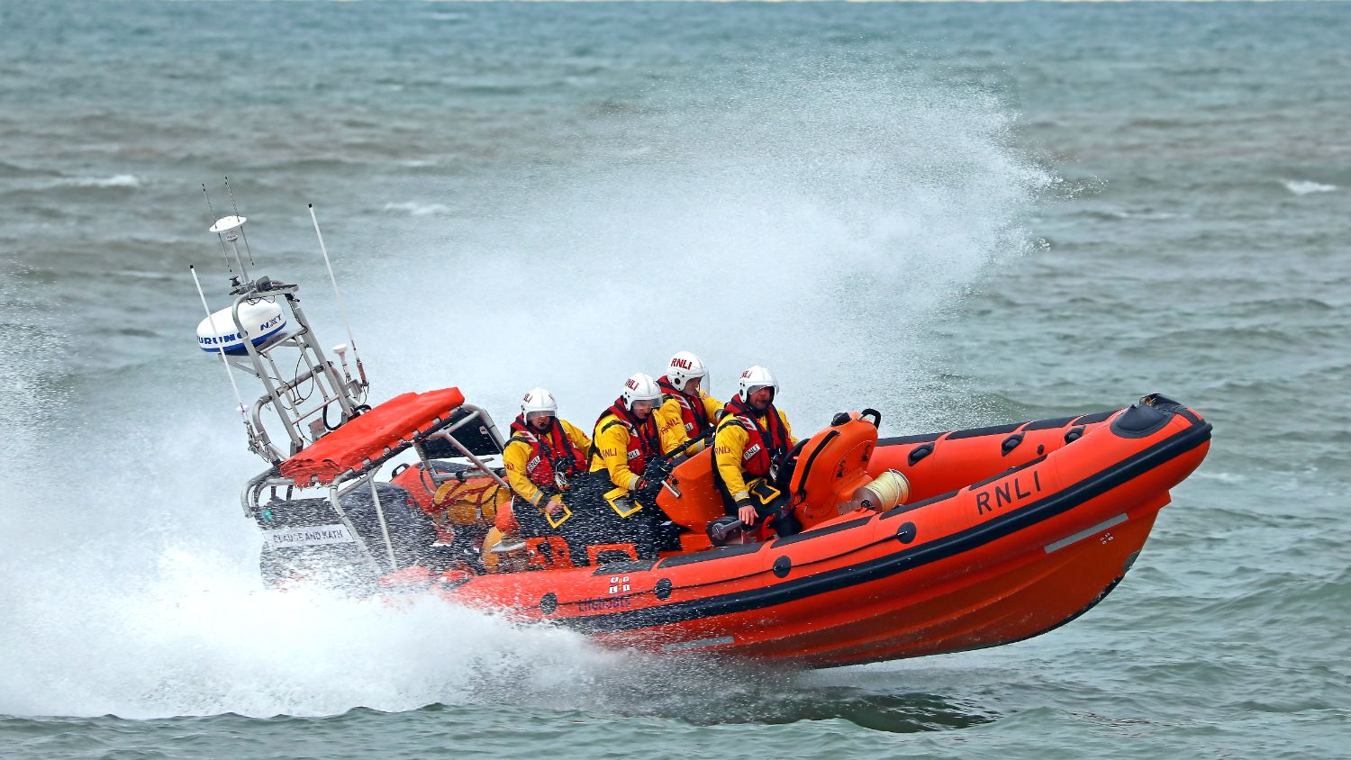 Orange inflatable RNLI lifeboat carrying several people in helmets and protective clothing, surrounded by large white spray as it moves through choppy water