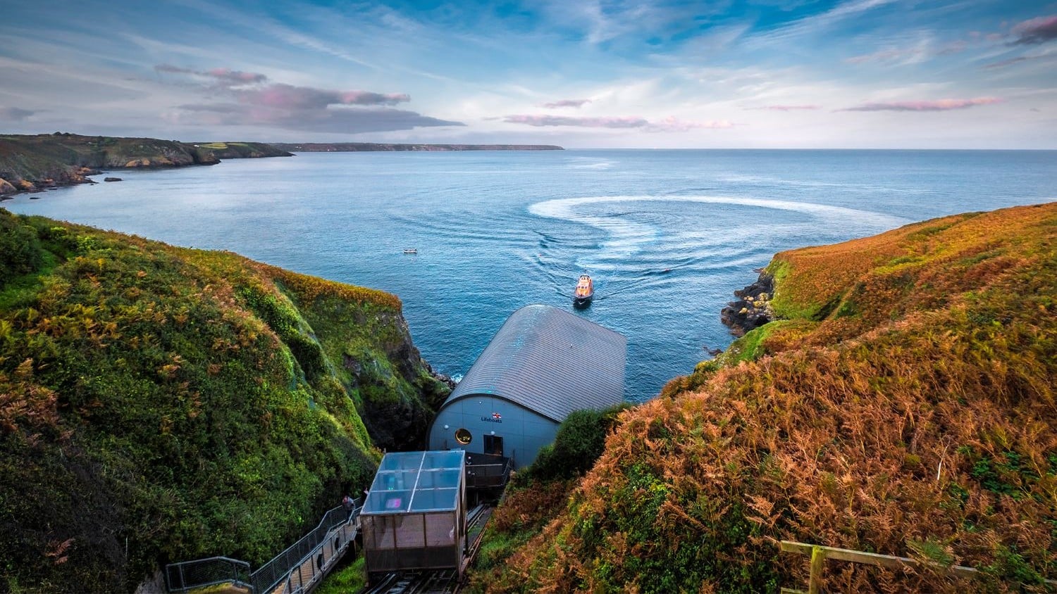 A building nestled between two hills looking out to see across a blue bay. A lifeboat in the bay has created a circular wake as it heads back to the station.