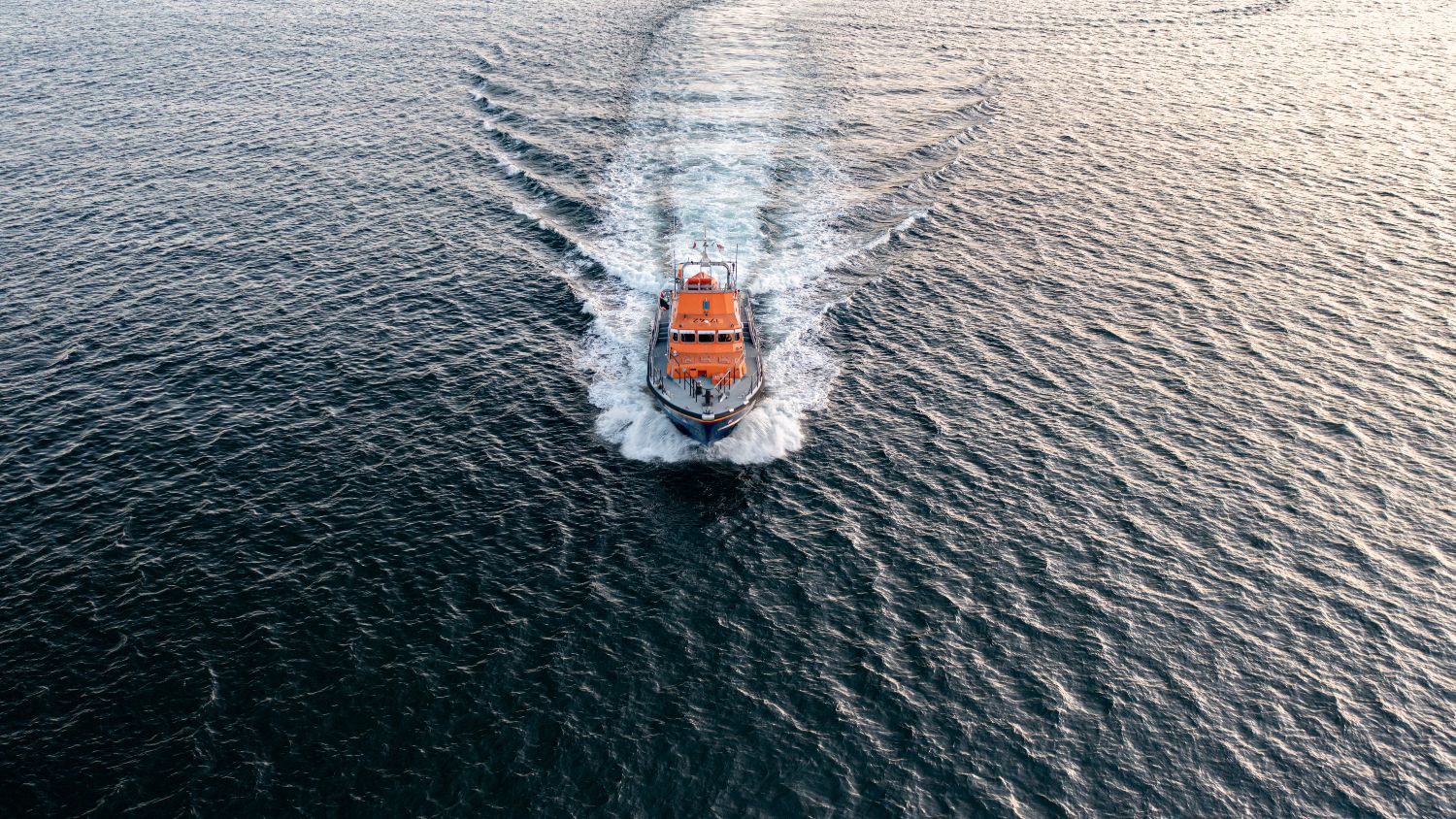 Aerial view of an orange RNLI lifeboat travelling away across dark, rippled water, leaving a wide V‑shaped white wake behind it.