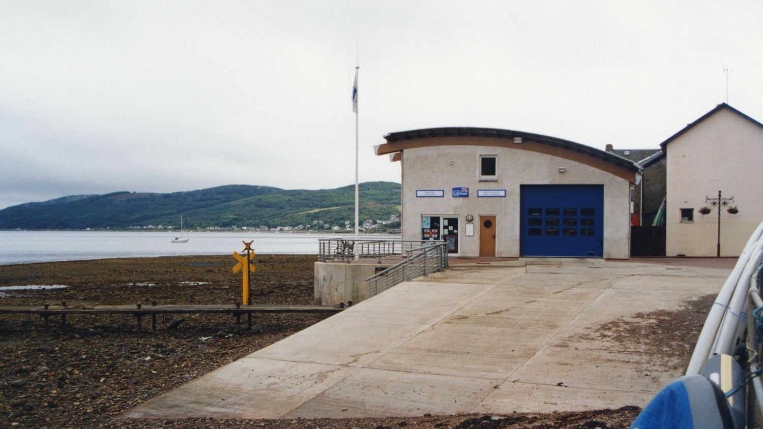 A grey building with curved roof and blue boat shed door on a shingle foreshore.