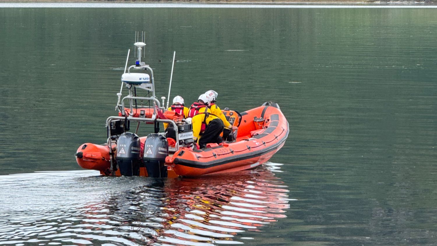 Orange inflatable RNLI lifeboat on calm water, carrying three people in helmets and high‑visibility jackets, with twin outboard engines at the rear.
