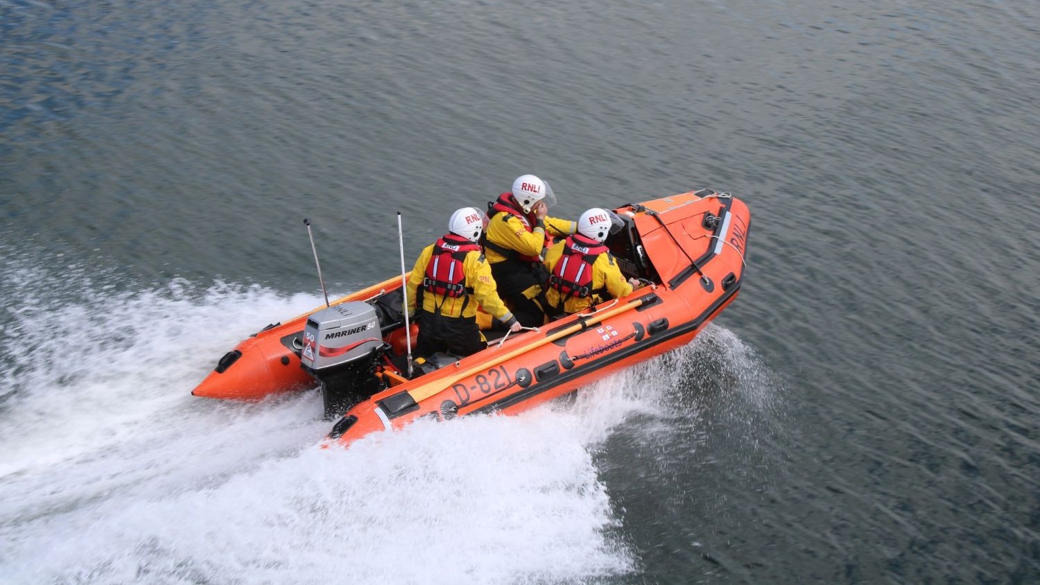 A small rib with three crew powers over calm grey water creating spray and a white wake.