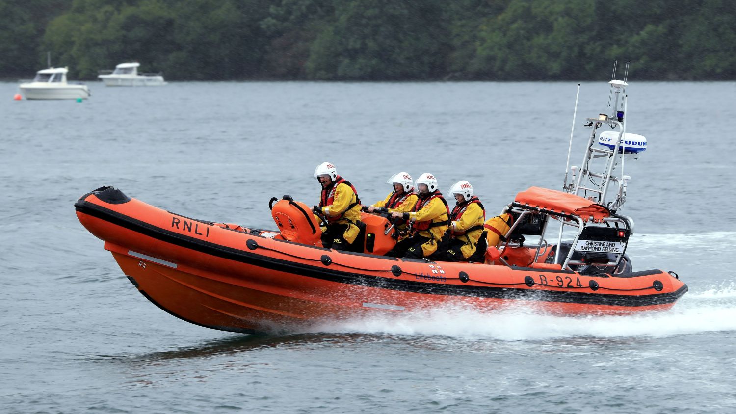 An orange RNLI lifeboat moves across the water carrying several crew members in yellow jackets and white helmets, under overcast skies with light ripples on the water.