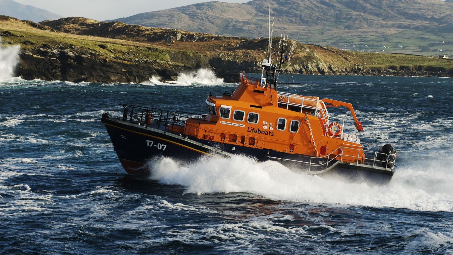 An orange RNLI lifeboat navigates choppy, dark blue water near a rocky coastline, with waves breaking and hills in the background under partly cloudy skies.