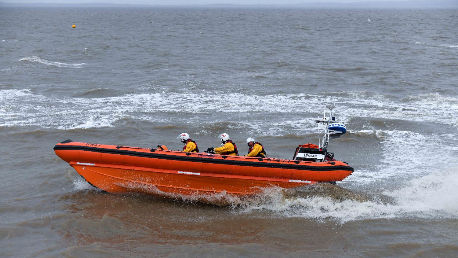 Orange inflatable RNLI lifeboat moving near the shoreline, with small waves breaking against the hull and three helmeted crew onboard.