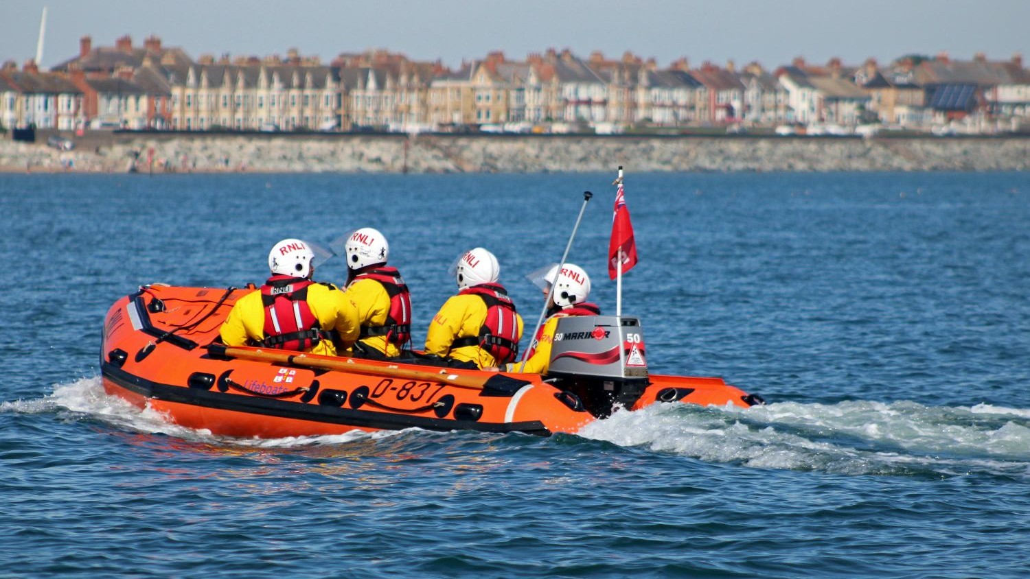 A small rib with four crew heading towards a town on calm blue seas.