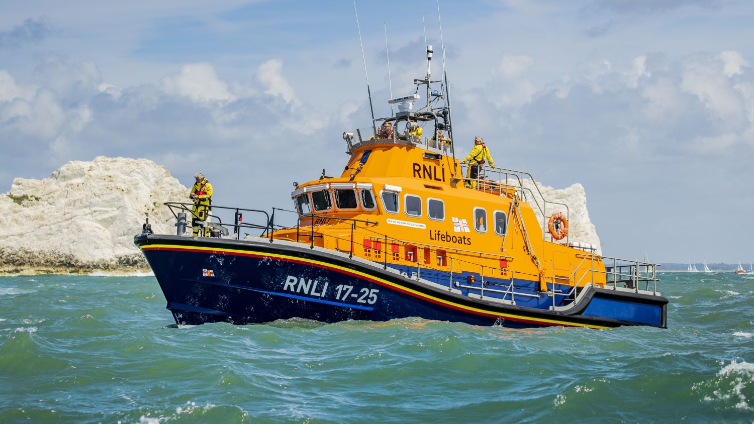An orange RNLI lifeboat in green choppy water near white cliffs, with crew members on deck under bright, partly cloudy skies.