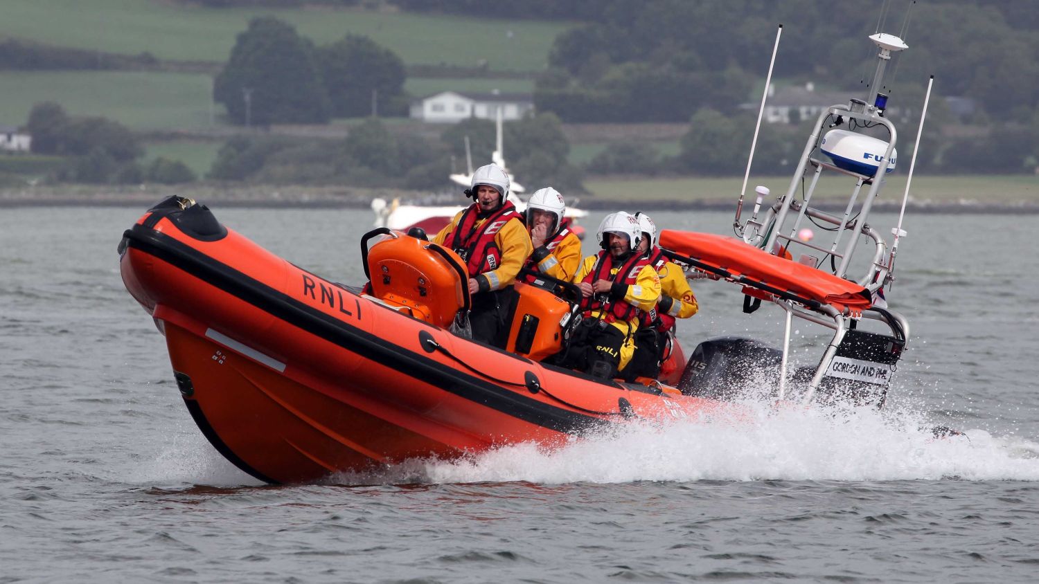 An orange RNLI lifeboat moves across calm grey water with several crew members on board in yellow jackets and helmets, with a green hillside and overcast sky in the background.