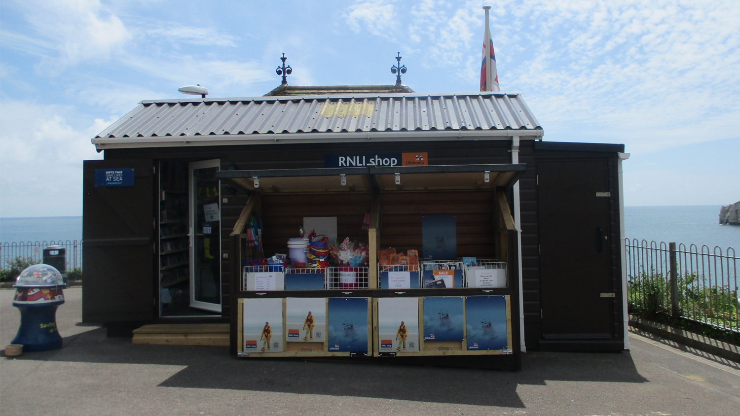 A small brown, wooden fronted shop on the top of a hill overlooking a blue sea.