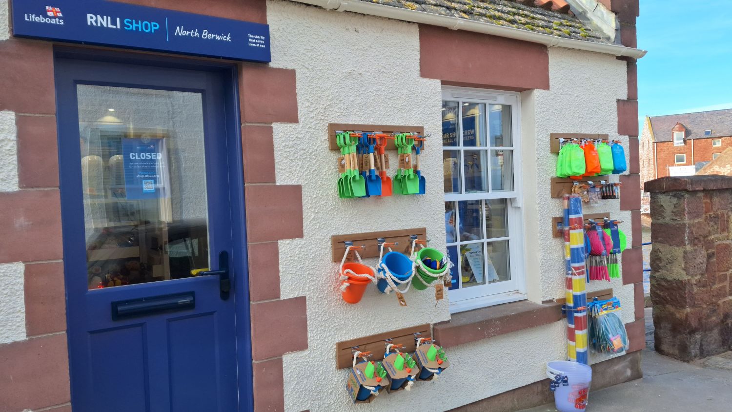 The front door of the North Berwick RNLI shop. The sun is shining and plastic buckets and spades adorn the front wall. 