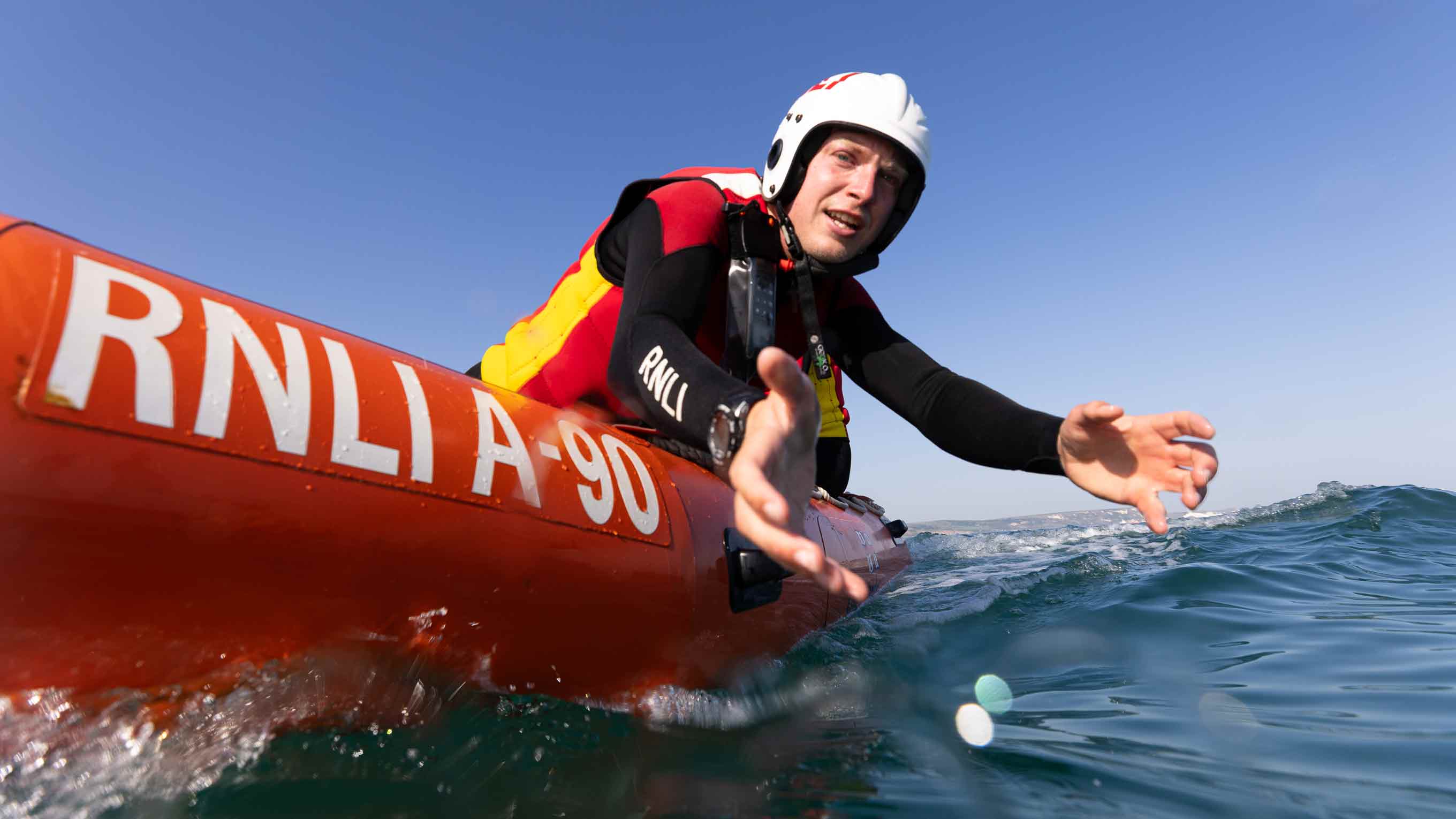 RNLI beach lifeguard Tom Allabarton reaches over the side of an inshore rescue boat to grab a casualty. He is wearing a white RNLI helmet and red and black lifeguard wetsuit. It's a bright sunny day with clear skies and blue seas.