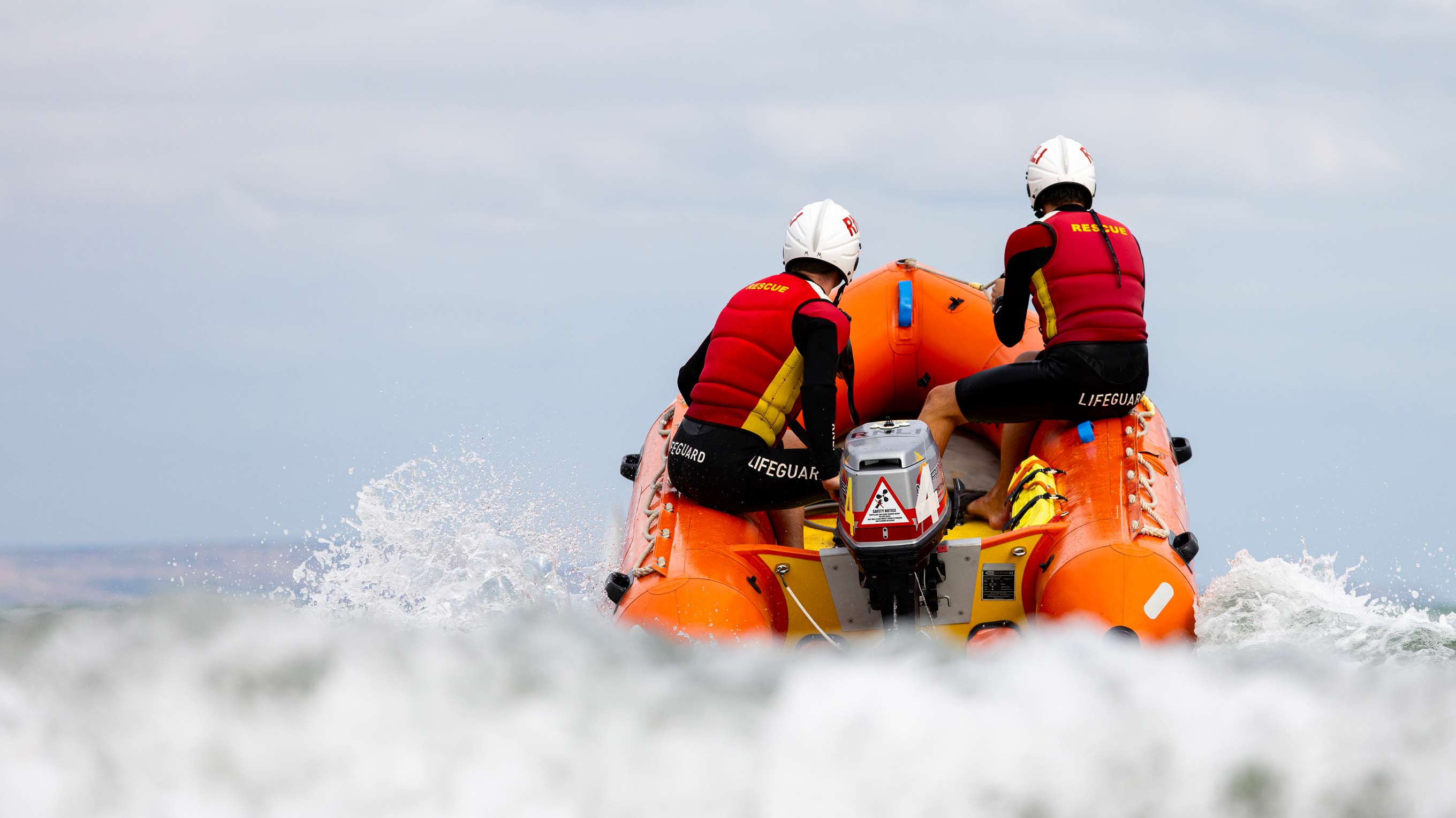 Two RNLI lifeguards on an Inshore Rescue Boat break through the crest of a wave on a cloudy day
